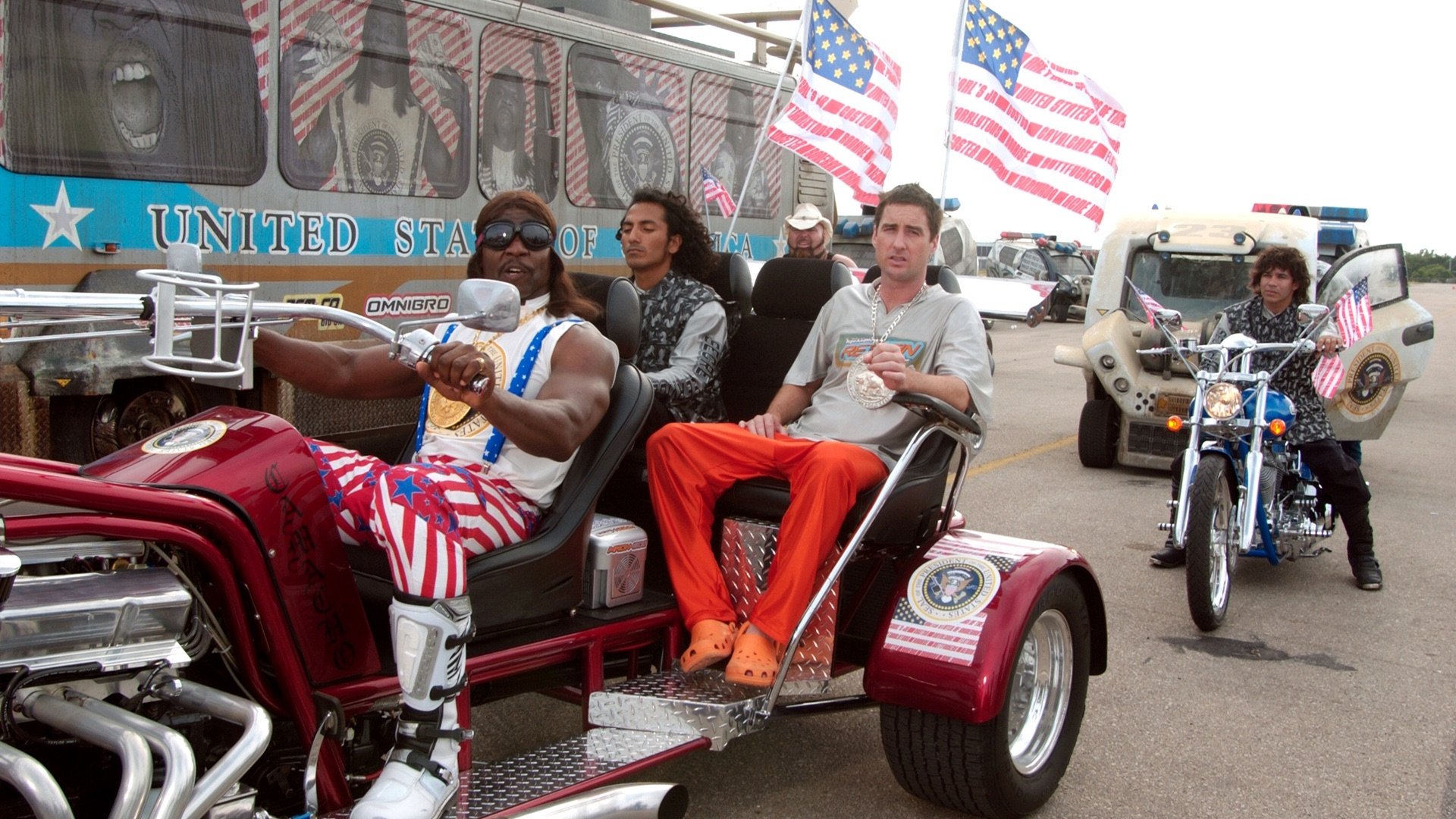 President Camacho rides the presidential trike flanked by American flags, Joe beside him in an orange prison jumpsuit (film still)